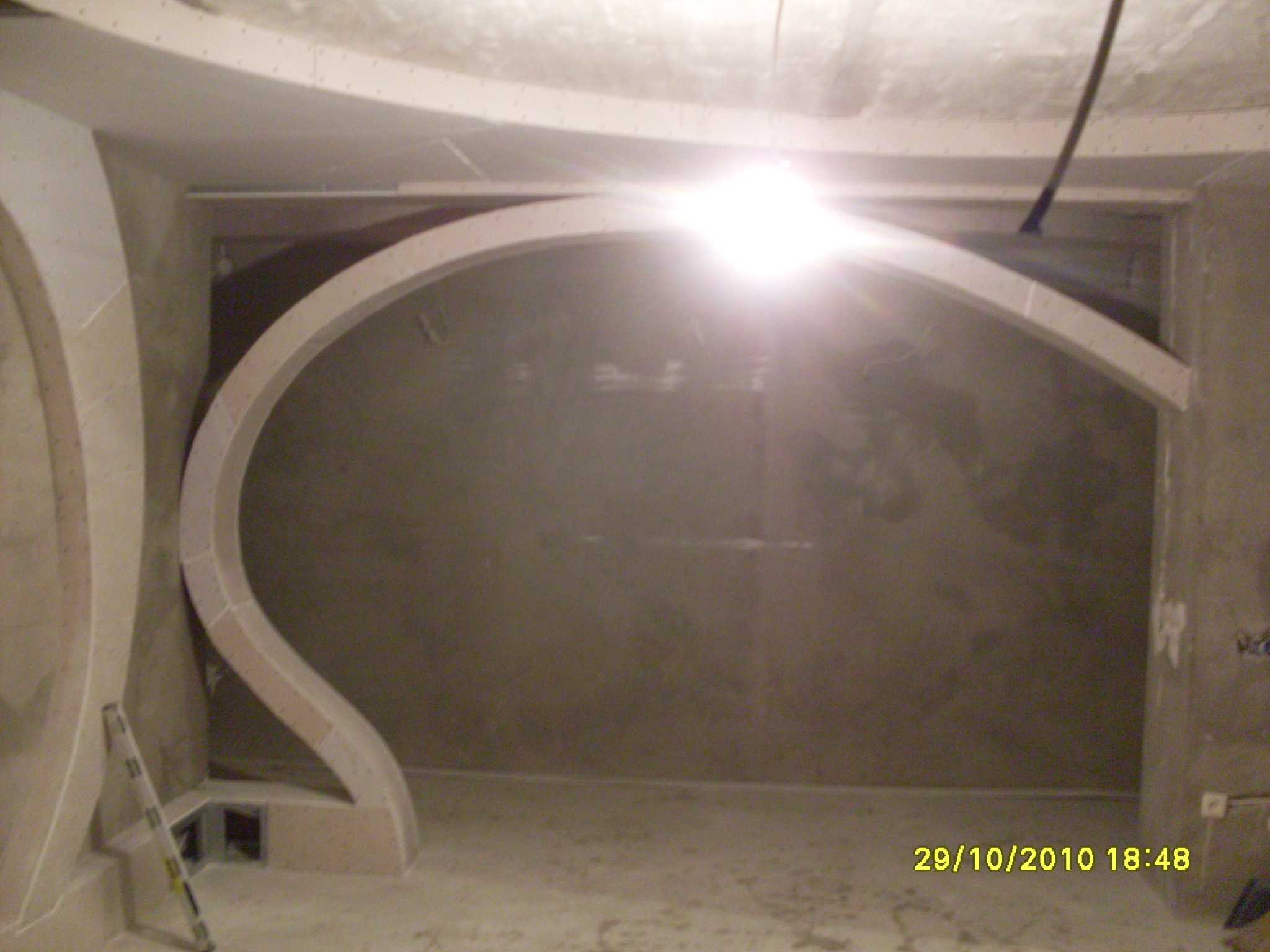 Niedokończona, zaokrąglona konstrukcja z płyt kartonowo-gipsowych na ścianie w trakcie prac wykończeniowych, widoczna drabina i surowy beton na ścianach.