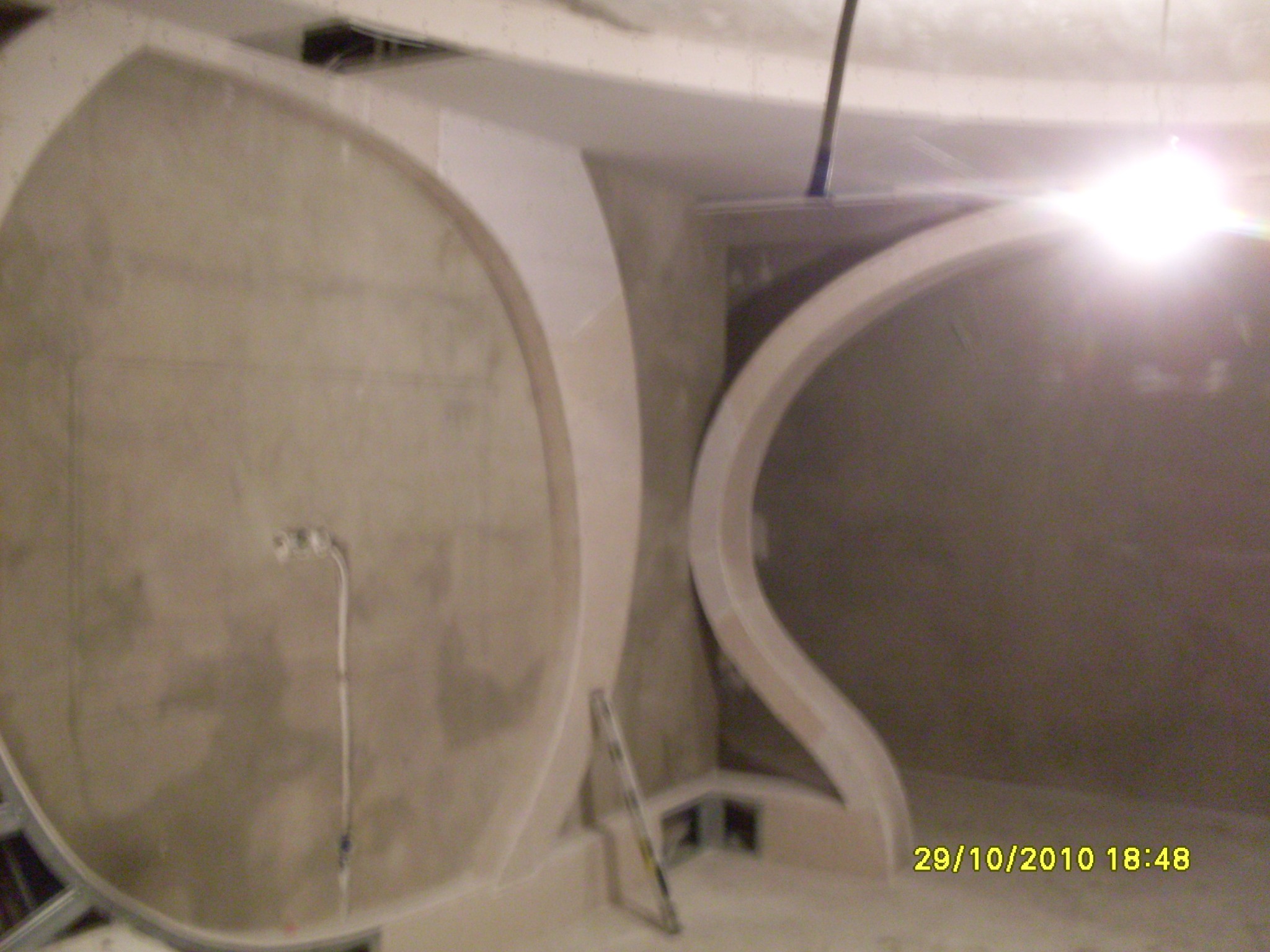 Niedokończona aranżacja łazienki z zaokrąglonymi ścianami i przygotowanymi instalacjami, widoczna drabina malarska i surowy stan wykończenia.