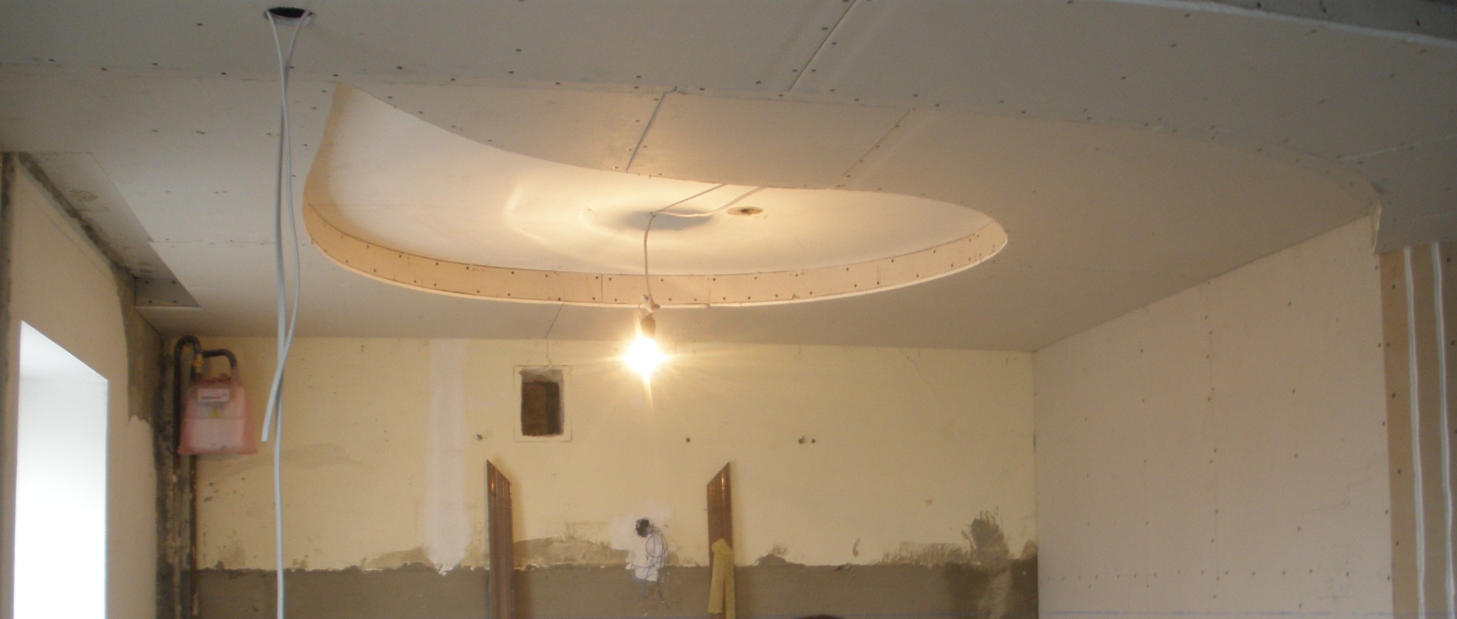 Niedokończony sufit podwieszany z płyt gipsowo-kartonowych z okrągłym elementem dekoracyjnym, widoczne przewody, ślady po pracach na ścianach, surowy stan wykończenia wnętrza.