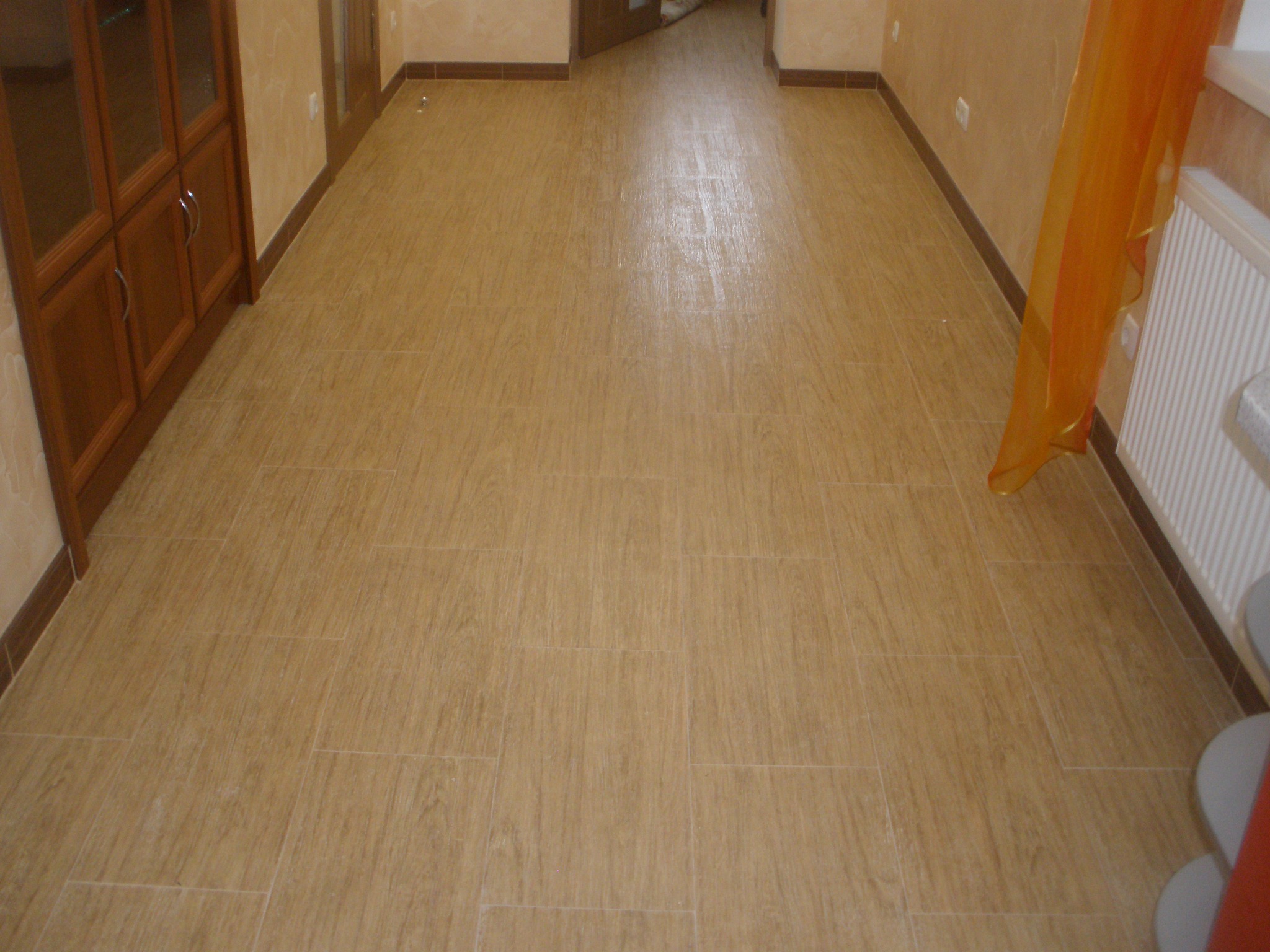 Beżowa podłoga z płytek imitujących drewno w jasnym wnętrzu, widoczna szafka z przeszkleniami i fragment pomarańczowej zasłony.