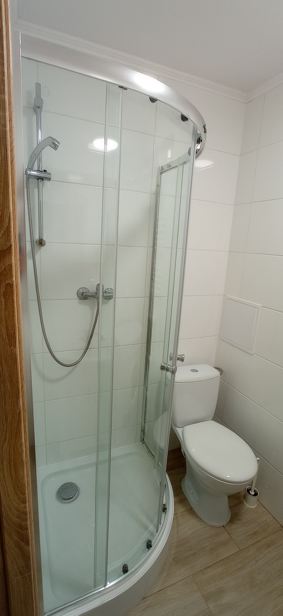 Nowoczesna kabina prysznicowa z otwartymi drzwiami, biała ceramika łazienkowa i jasne płytki imitujące drewno na podłodze.