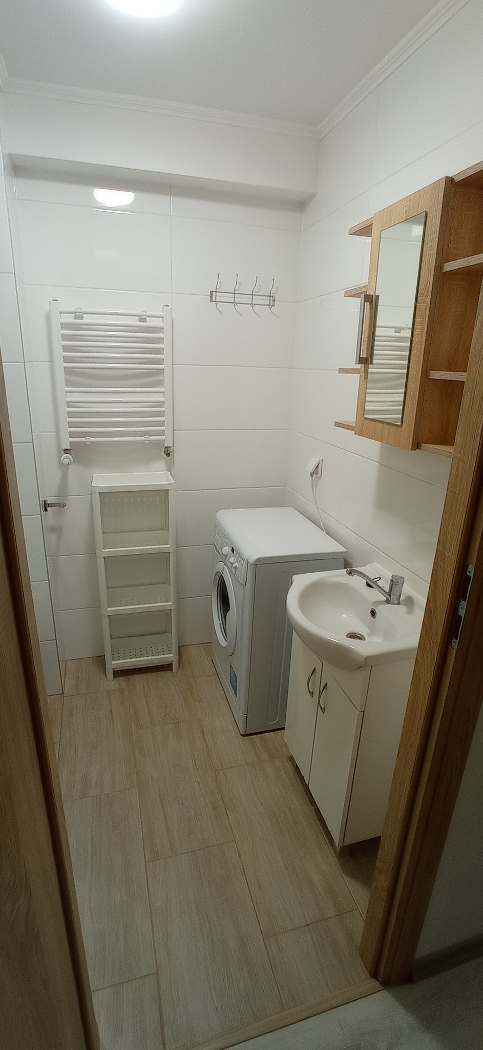 Jasna łazienka po remoncie z białymi płytkami, pralką, umywalką z szafką, grzejnikiem drabinkowym i drewnianą szafką z lustrem