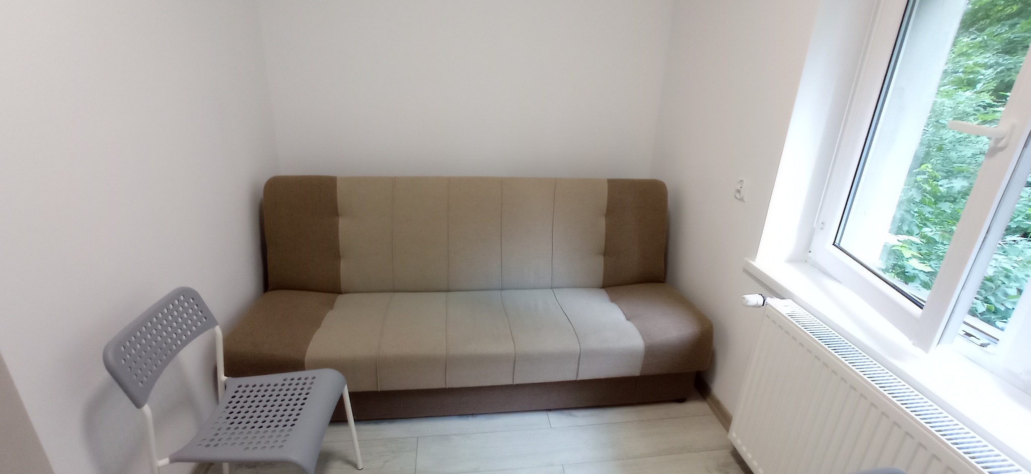 Pokój z beżową sofą, szarym krzesłem i białym grzejnikiem pod oknem z widokiem na zieleń.