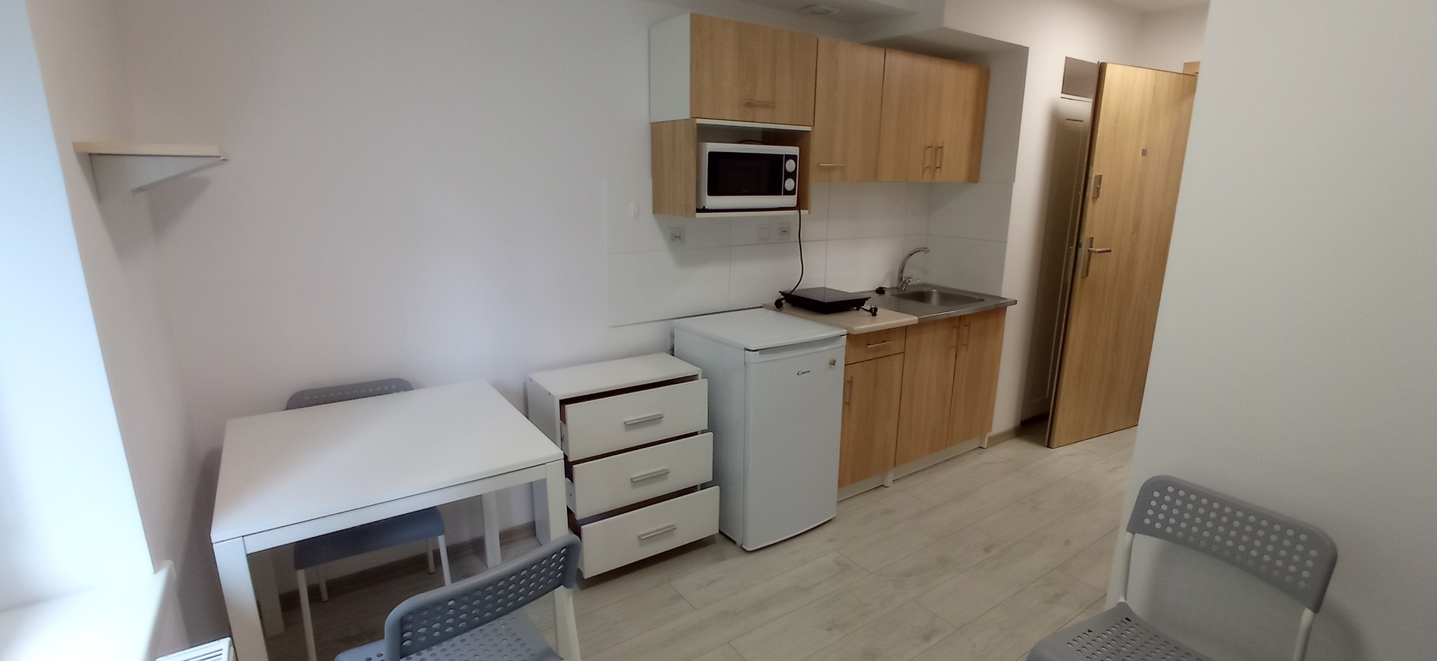 Widok na małą kuchnię w kawalerce z białymi ścianami, jasnymi drewnianymi szafkami, białym sprzętem AGD i prostym białym stołem z szarymi krzesłami.