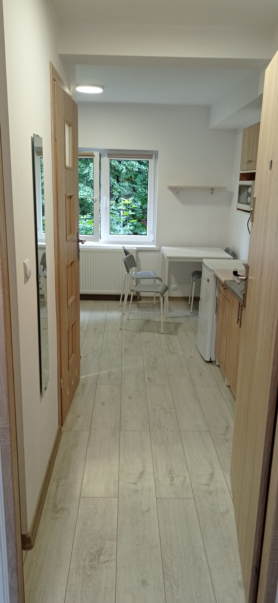 Jasne, świeżo wykończone wnętrze kawalerki z podłogą imitującą drewno, widok z przedpokoju na aneks kuchenny z białymi meblami i stolikiem przy oknie z widokiem na zieleń.