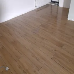 Ułożona podłoga z płytek imitujących drewno w jasnym pomieszczeniu z białymi ścianami i grzejnikiem; widoczny fragment przewodu elektrycznego wystającego z podłogi.