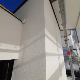 Świeżo nałożony, biały tynk strukturalny na ścianie budynku, widoczne rusztowanie i fragment niebieskiego nieba.