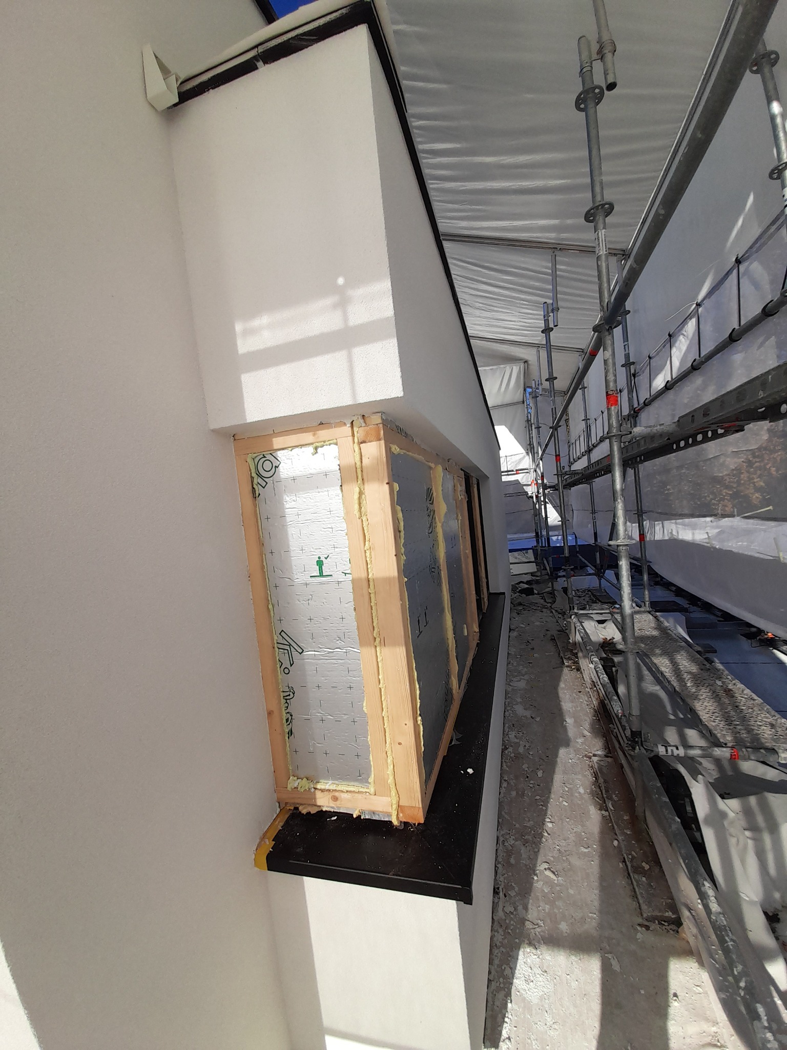 Montaż okna z drewnianą ramą i izolacją z folii aluminiowej, widoczne rusztowanie i przygotowana elewacja zewnętrzna budynku.