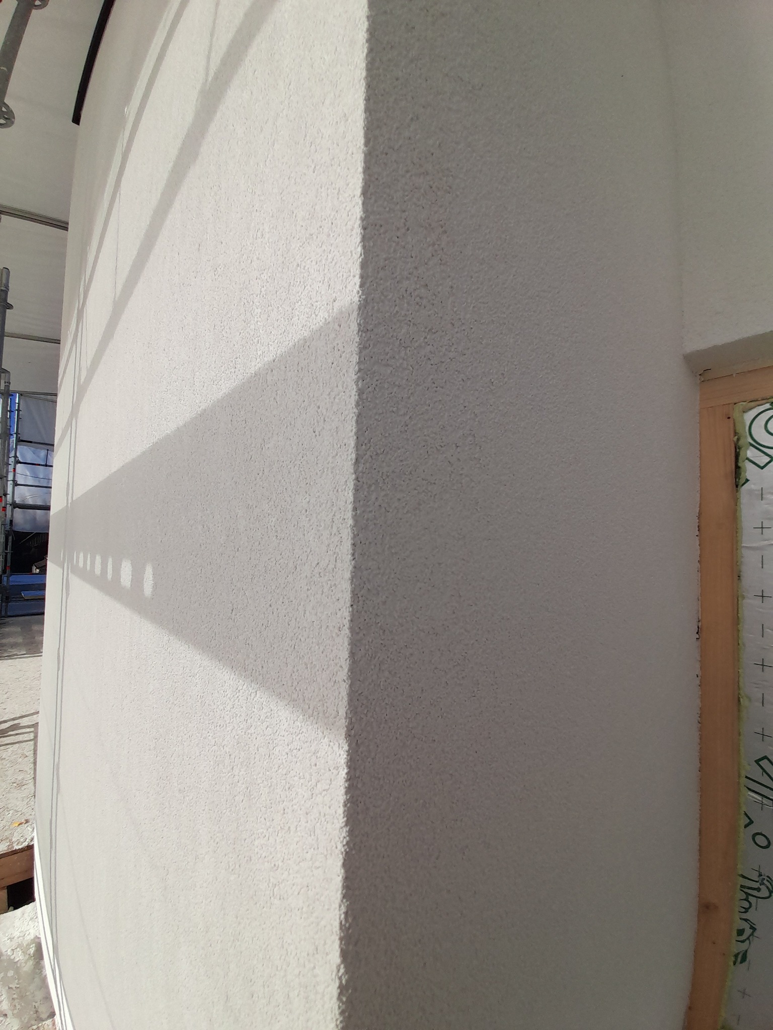 Świeżo nałożony jasny tynk strukturalny na rogu budynku, z widocznym cieniem rusztowania i fragmentem okna z izolacją.