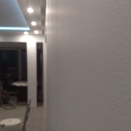 Pakerman - Świeżo pomalowana, gładka ściana w odcieniach szarości, widoczna faktura, w tle fragment pomieszczenia z oświetleniem sufitowym i krzesłami.