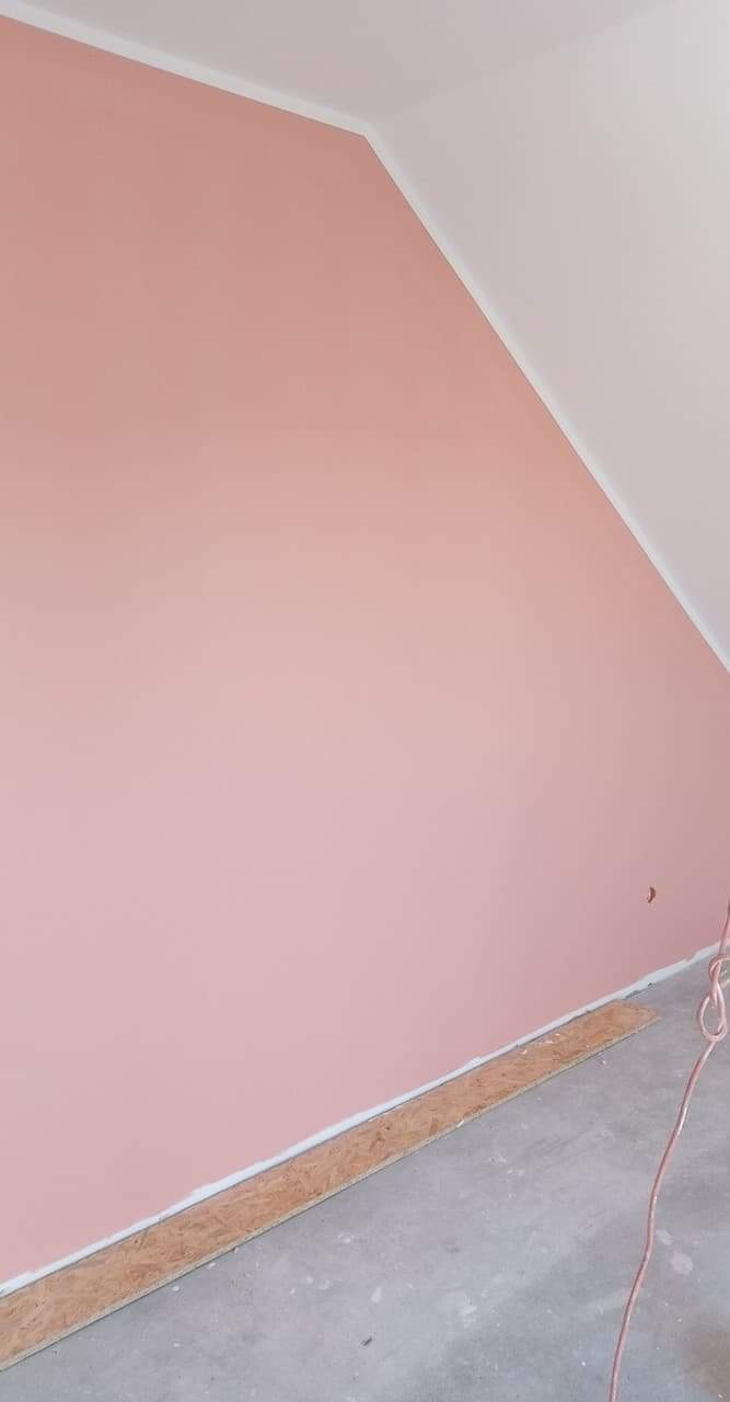 Ściana w pokoju na poddaszu pomalowana na delikatny róż, z białym wykończeniem sufitu i listwami przypodłogowymi, zabezpieczona płytą OSB przy podłodze przed zabrudzeniem.