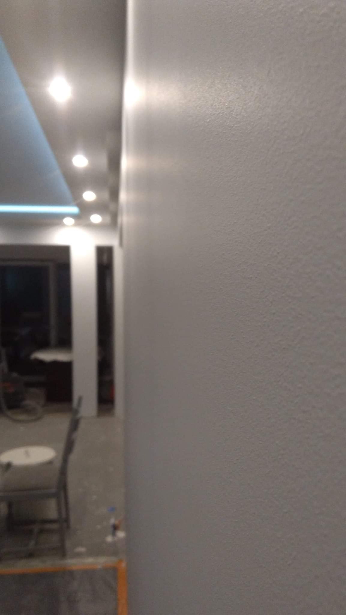 Świeżo pomalowana, gładka ściana w odcieniach szarości, widoczna faktura, w tle fragment pomieszczenia z oświetleniem sufitowym i krzesłami.