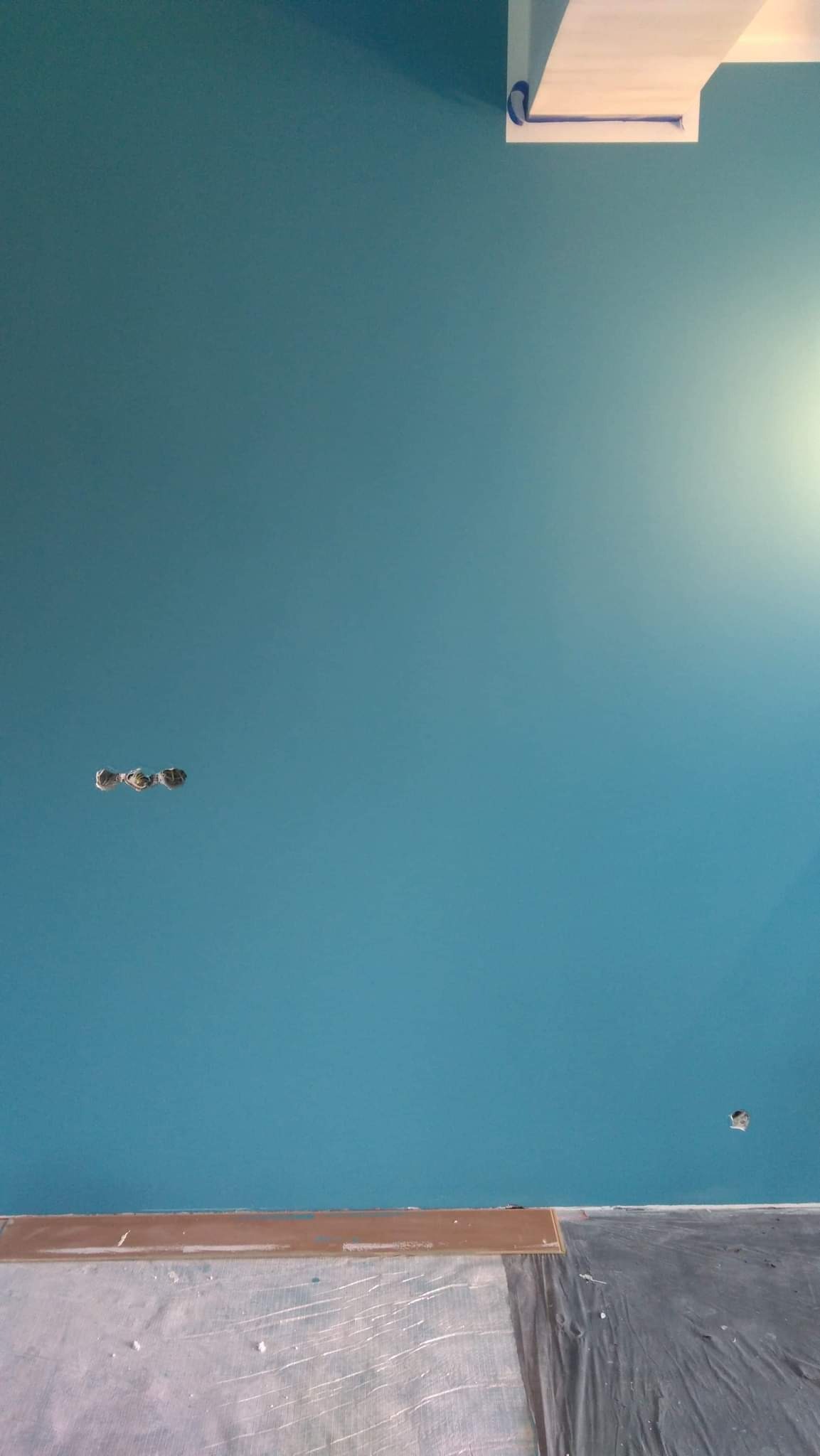 Świeżo pomalowana na turkusowo ściana z widocznymi trzema otworami na kable i pojedynczym otworem w prawym górnym rogu, zabezpieczona taśmą malarską przy suficie i folią ochronną na podłodze.