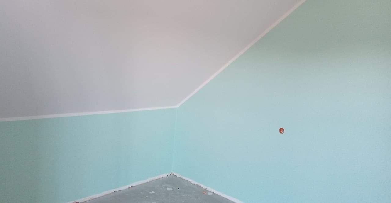 Świeżo pomalowane wnętrze poddasza z jasnymi, turkusowymi ścianami i białym skośnym sufitem, widoczne gniazdko elektryczne w ścianie.