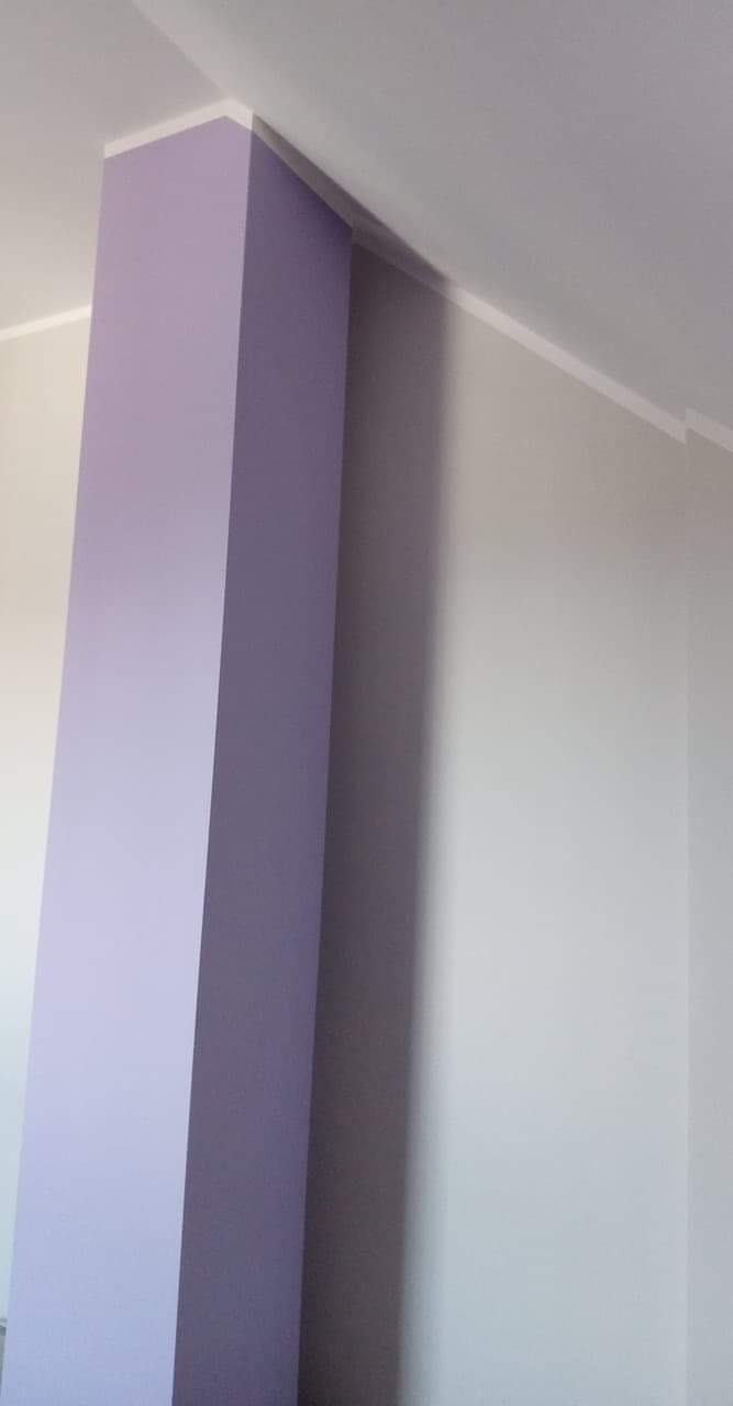 Pomalowany na liliowo filar narożny w pomieszczeniu z szarą ścianą i białym sufitem, wykończony białymi listwami.