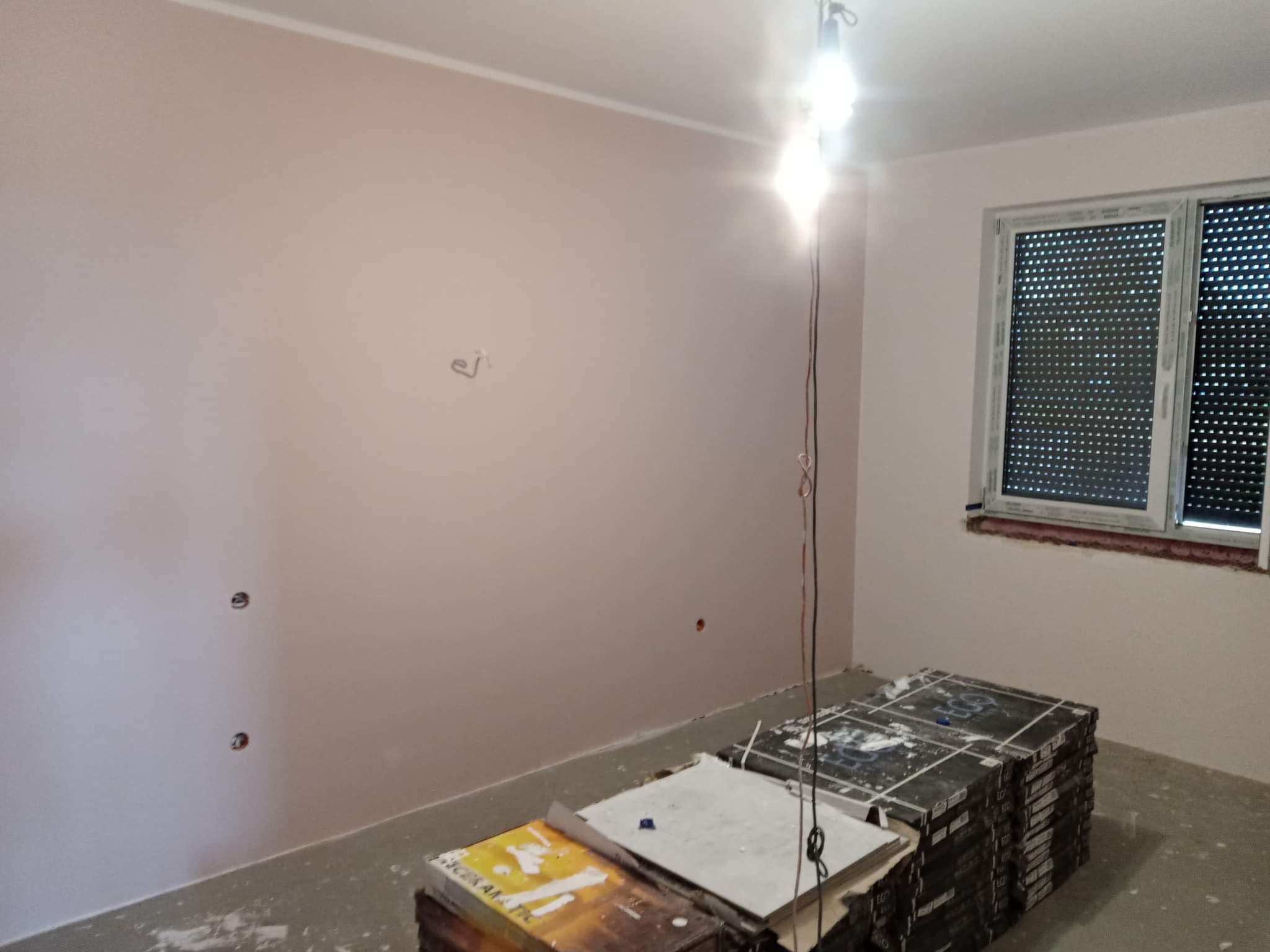 Pomieszczenie po świeżym pomalowaniu na delikatny róż, z widocznymi puszkami elektrycznymi i stertą płytek na podłodze, oświetlone jasną żarówką budowlaną.