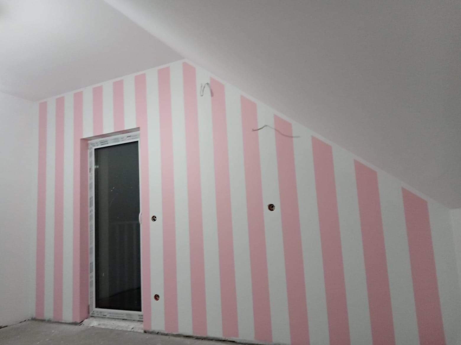 Pokój na poddaszu z ukończonymi ścianami w różowo-białe pionowe pasy, widoczne puszki elektryczne bez osprzętu i wystające przewody, okno balkonowe w białej ramie.