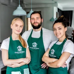 Trzyosobowy zespół sprzątający Clean Republic w zielonych fartuchach z logo, stojący w nowoczesnym wnętrzu z rękawicami i ściereczką w kieszeni, uśmiechnięty do kamery.