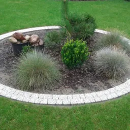 Okrągła rabata z białej kostki granitowej otoczona trawnikiem, wypełniona korą, ozdobnymi trawami i niskimi krzewami, z doniczkami z kamieniami w tle.