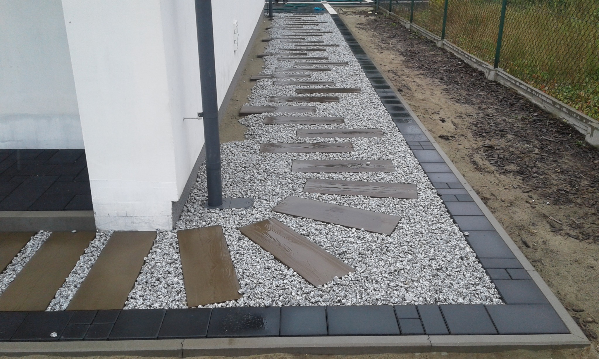 Ścieżka z płyt imitujących drewno ułożona na jasnym żwirze, obrzeża z ciemnej kostki brukowej przy białej ścianie budynku i metalowym słupie