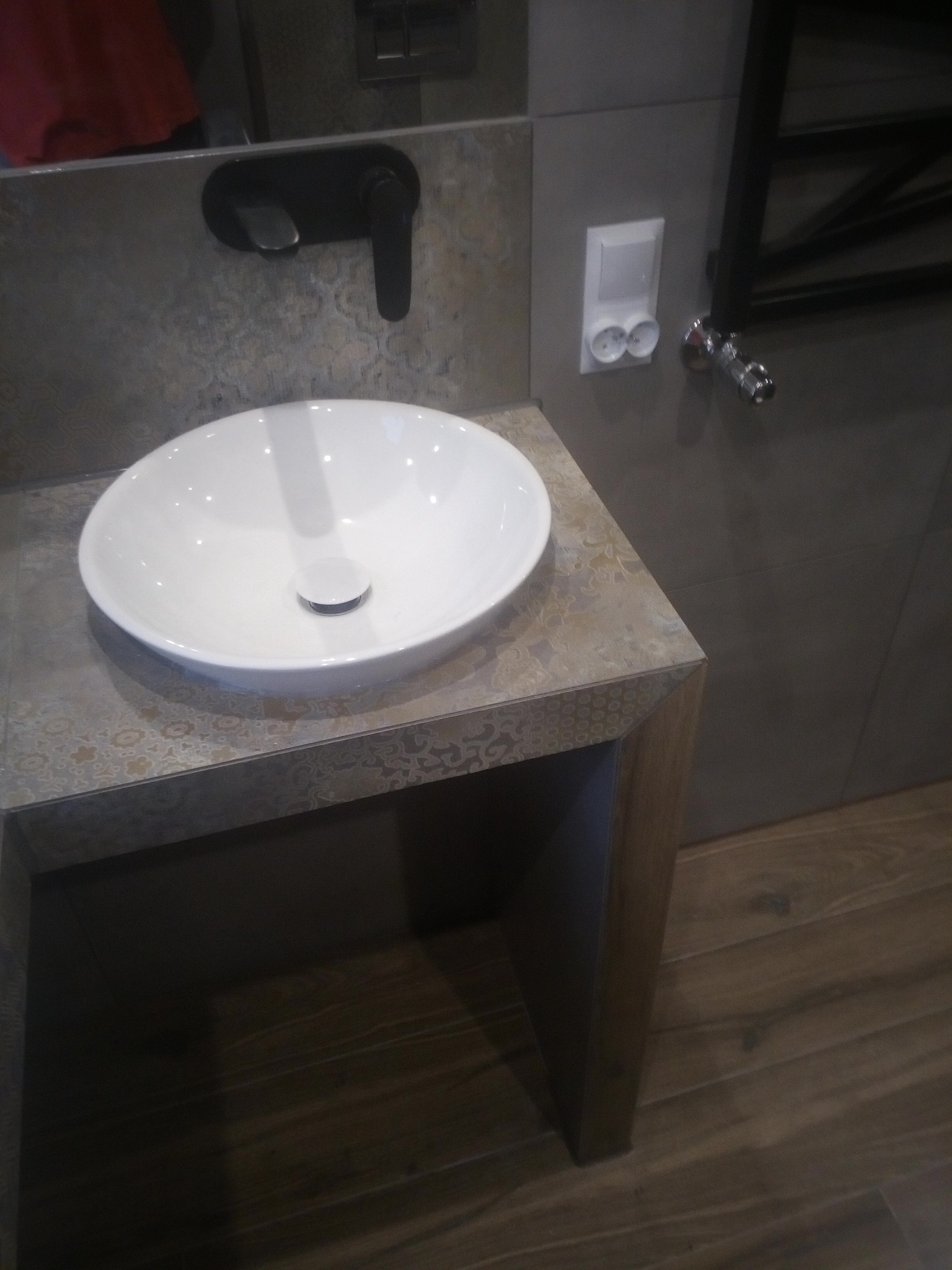 Biała, owalna umywalka nablatowa z czarną baterią, zamontowana na szafce z blatem w beżowo-szarym odcieniu z delikatnym wzorem, na tle szarych płytek i drewnianej podłogi.