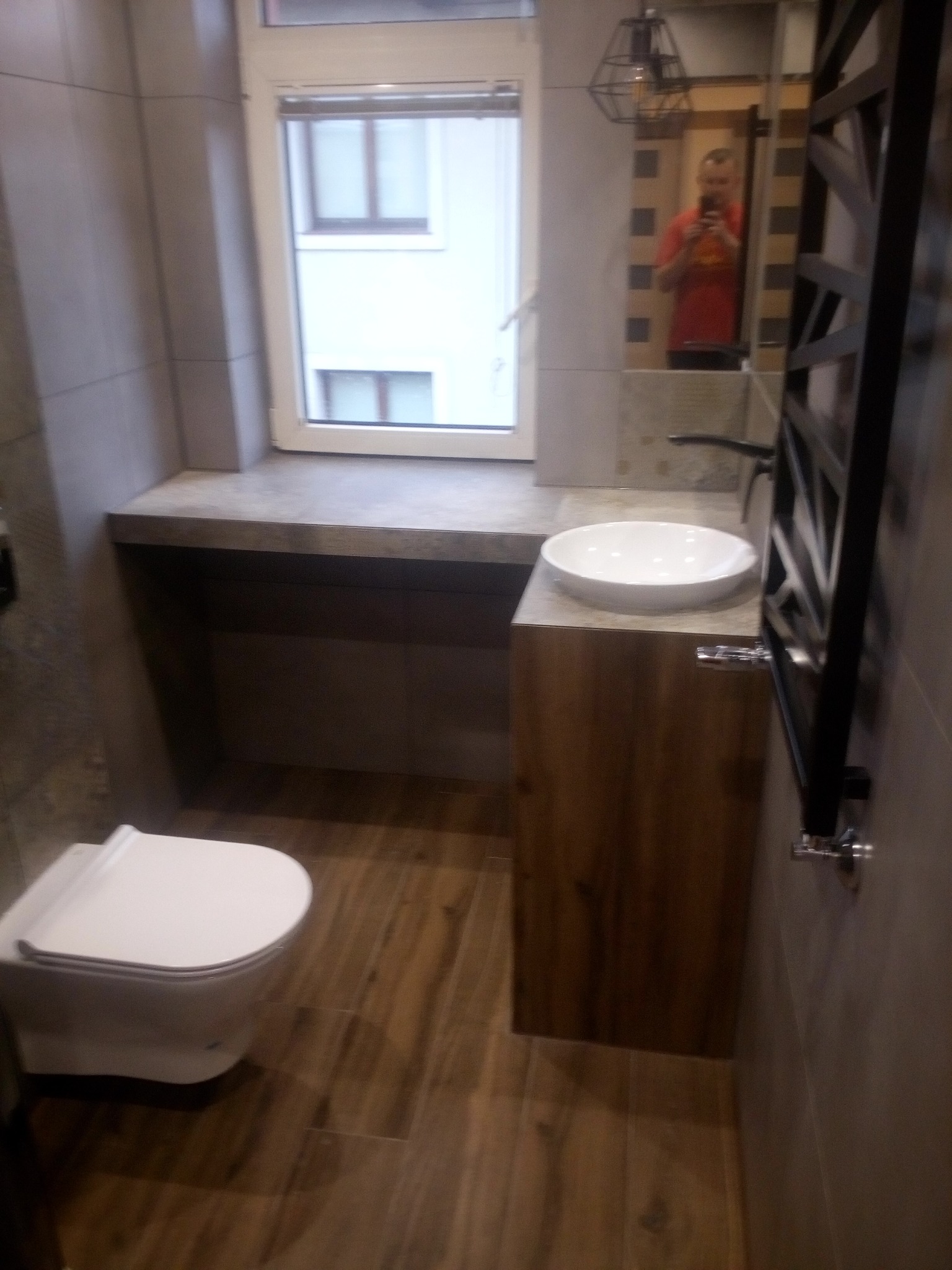 Nowoczesna łazienka z drewnopodobną podłogą, wiszącą toaletą, umywalką nablatową na drewnianej szafce i grzejnikiem drabinkowym; w lustrze odbija się osoba robiąca zdjęcie.