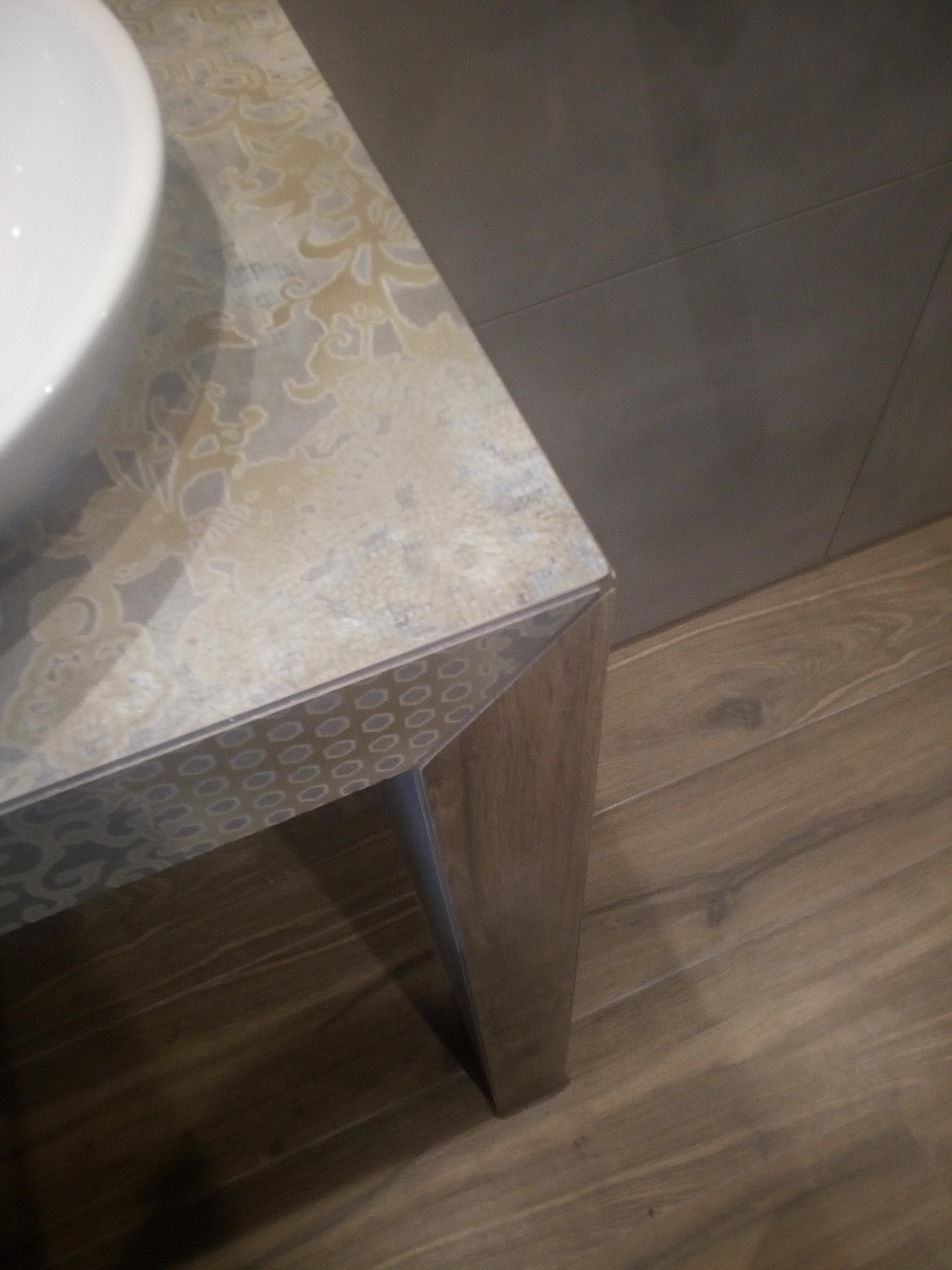 Blat umywalki z wzorem paisley i zdobioną krawędzią, wsparty na drewnianej nodze, na tle szarej ściany i drewnopodobnej podłogi.