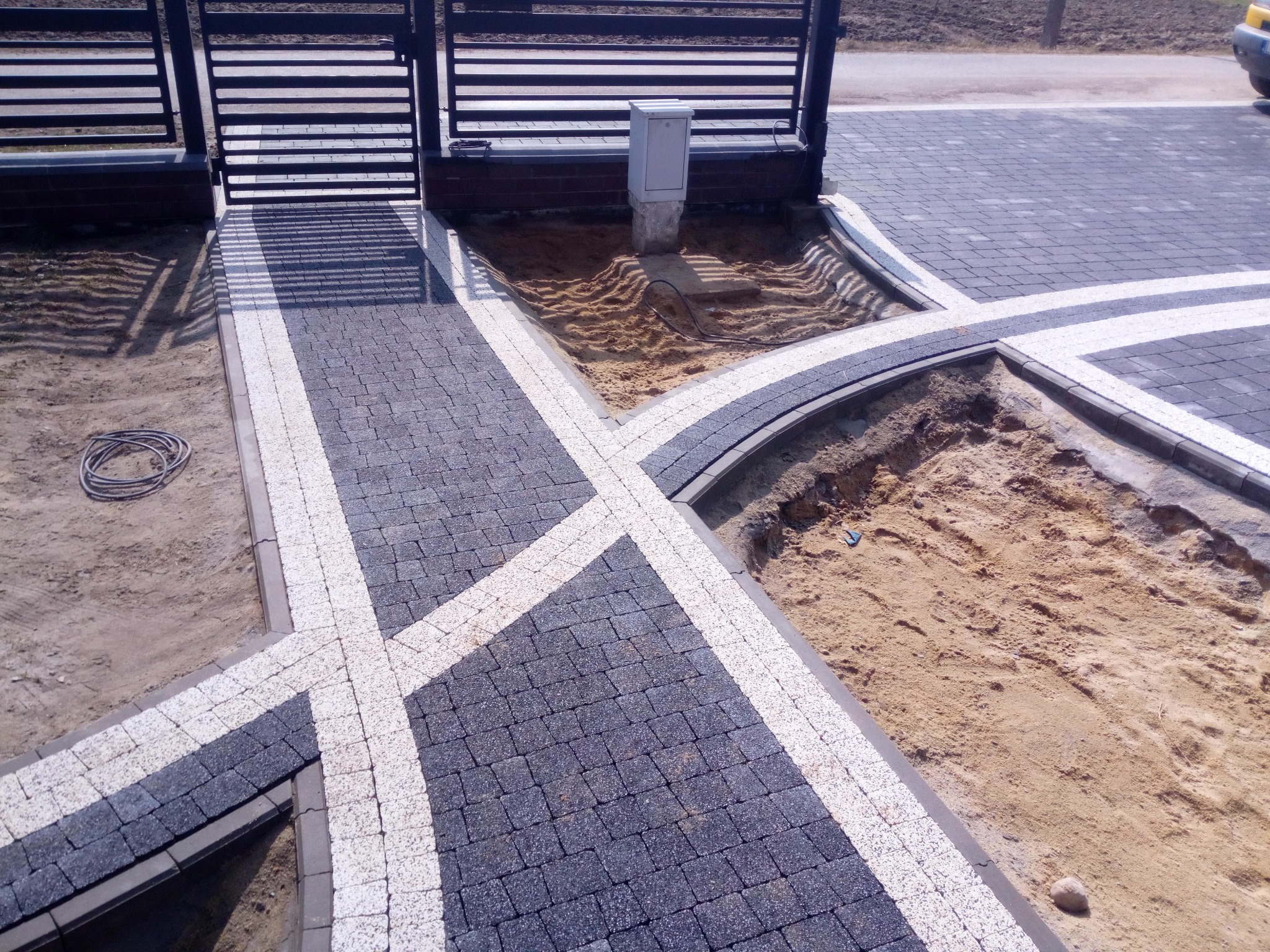 Fragment podjazdu z kostki brukowej w kolorze grafitu z białymi akcentami, widoczne przygotowane podłoże pod trawnik i brama wjazdowa, kabel elektryczny leżący obok.