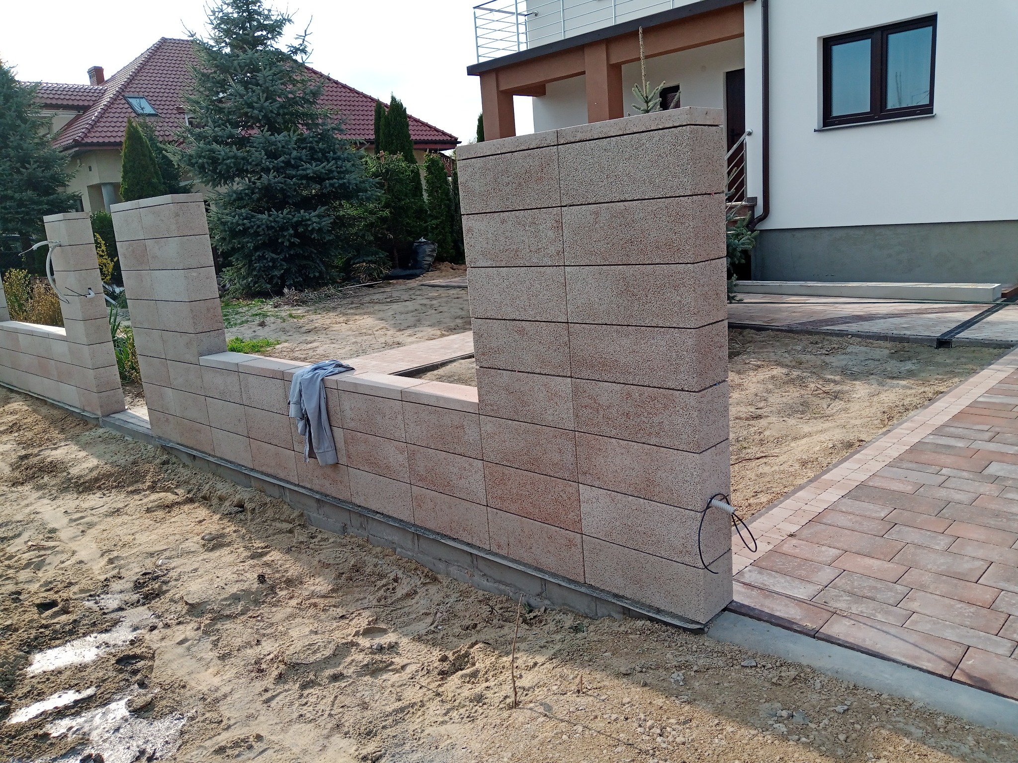 Wznoszenie ogrodzenia z bloczków betonowych w kolorze piaskowym, z widoczną konstrukcją fundamentową i częściowo ułożoną kostką brukową obok, na tle domu jednorodzinnego z czerwonym dachem.