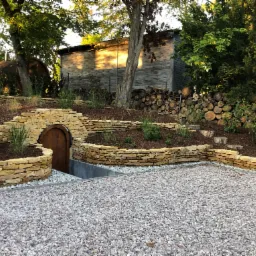 Ogród z kamiennymi murkami oporowymi, drewnianą piwniczką ziemną i żwirowym podłożem. W tle drewniany budynek i stos drewna.