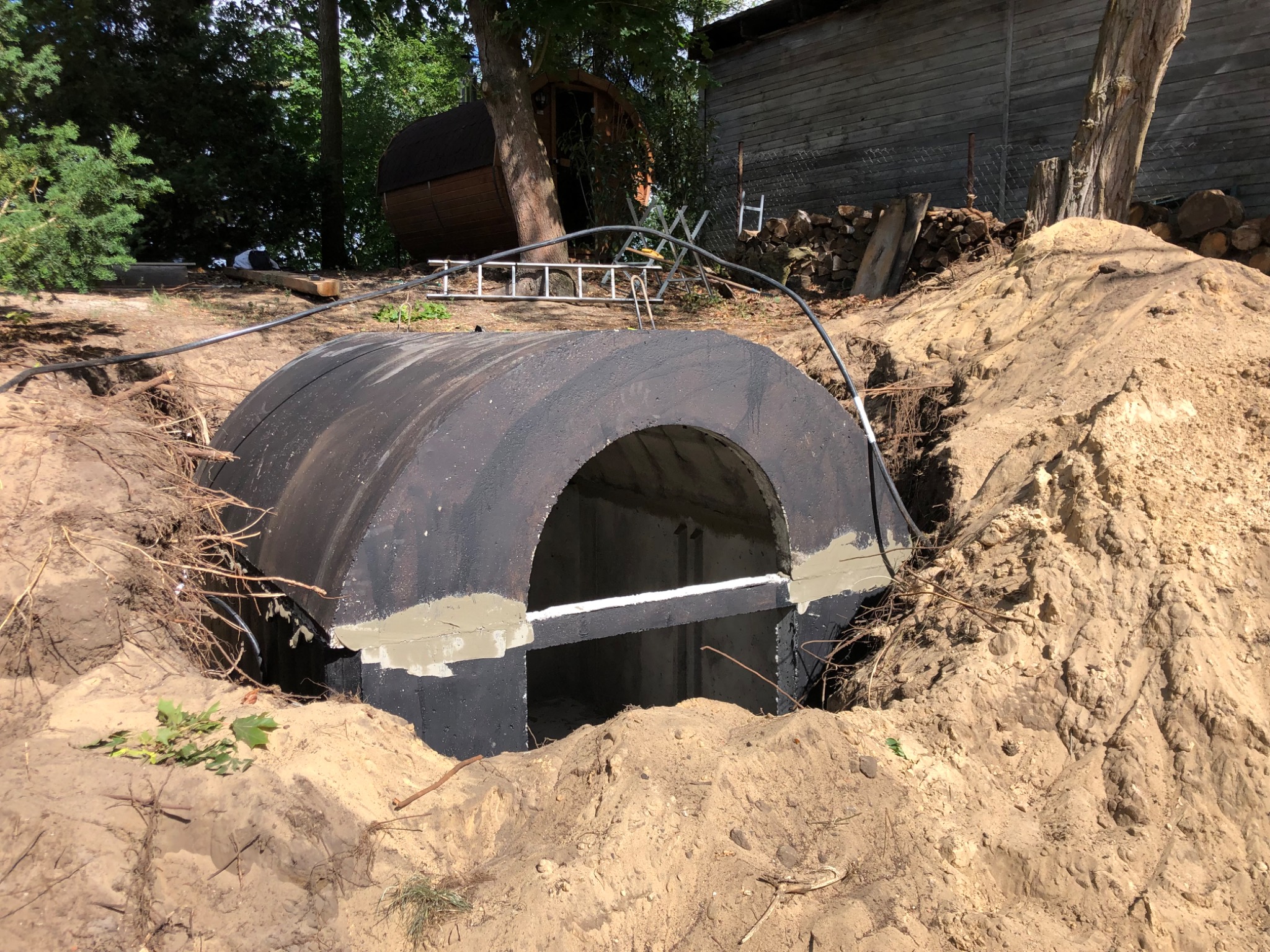 Częściowo zakopana, czarna, betonowa konstrukcja tunelowa z widocznymi śladami zaprawy, otoczona piaskiem i zielenią. W tle drabina, drewniany budynek i sauna.