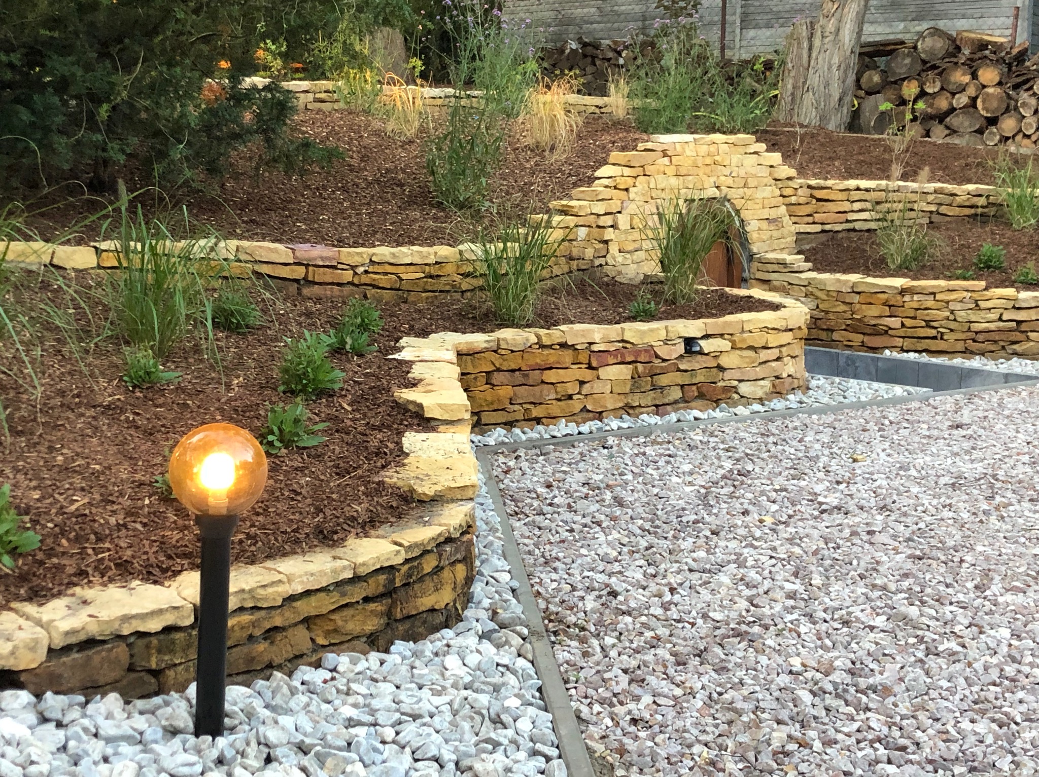 Ogród z kamiennymi murkami, roślinami i oświetleniem. Dekoracyjny tunel z kamienia. Ścieżka z białego żwiru. W tle ułożone drewno.