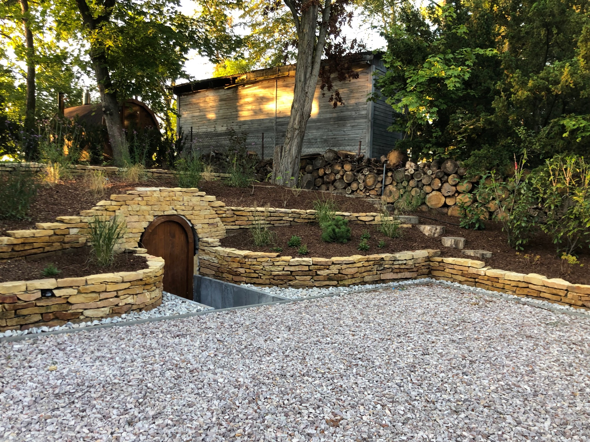 Ogród z kamiennymi murkami oporowymi, drewnianą piwniczką ziemną i żwirowym podłożem. W tle drewniany budynek i stos drewna.