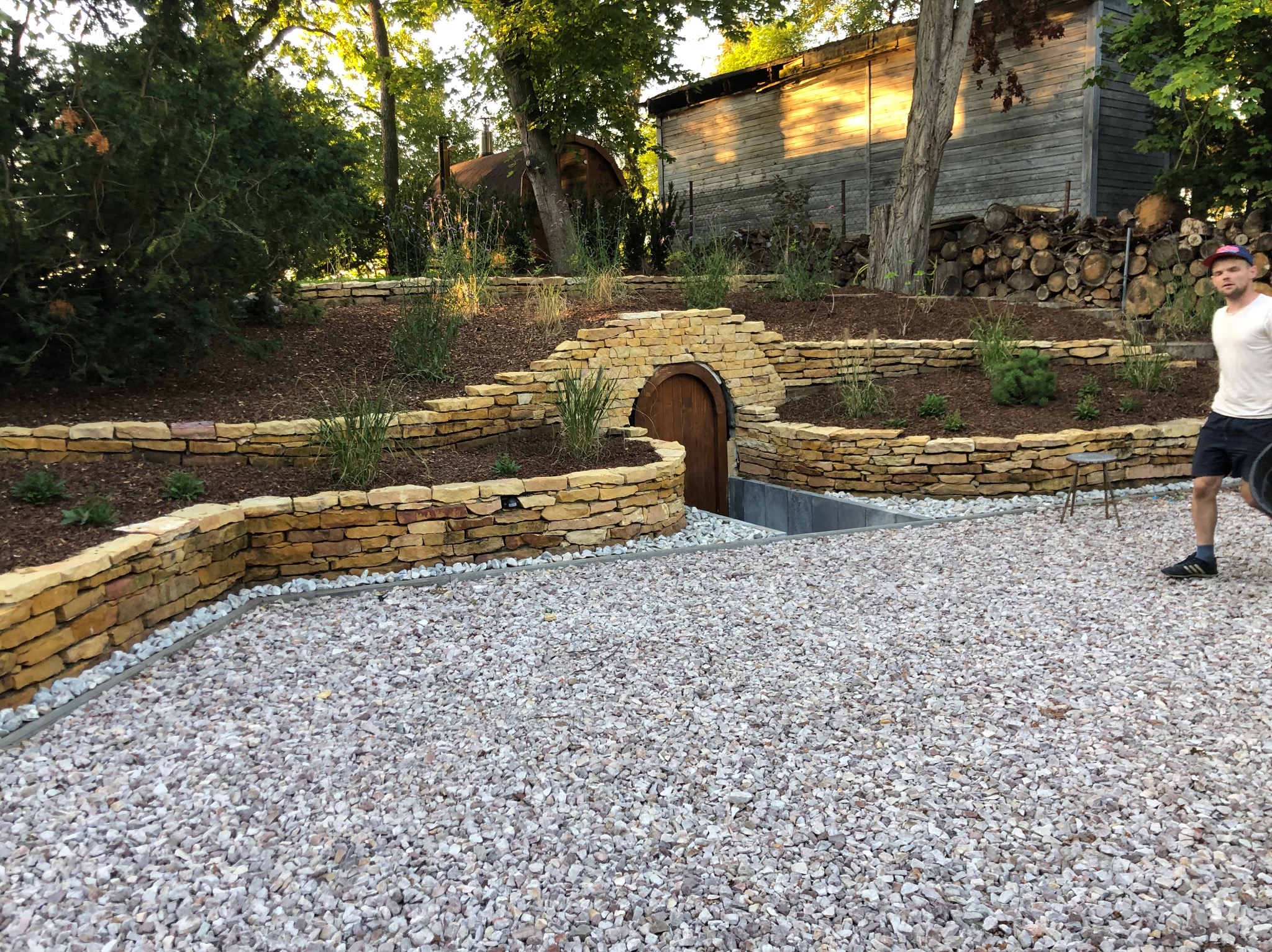 Ogród z kamiennymi murkami, drewnianymi drzwiami do piwnicy i wysypanym żwirem. W tle budynek gospodarczy i stos drewna. Osoba w kadrze.
