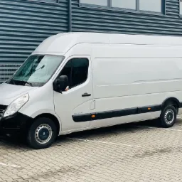 Srebrny samochód dostawczy Renault Master zaparkowany na parkingu z kostki brukowej, na tle szarej, metalowej ściany budynku z oknami.