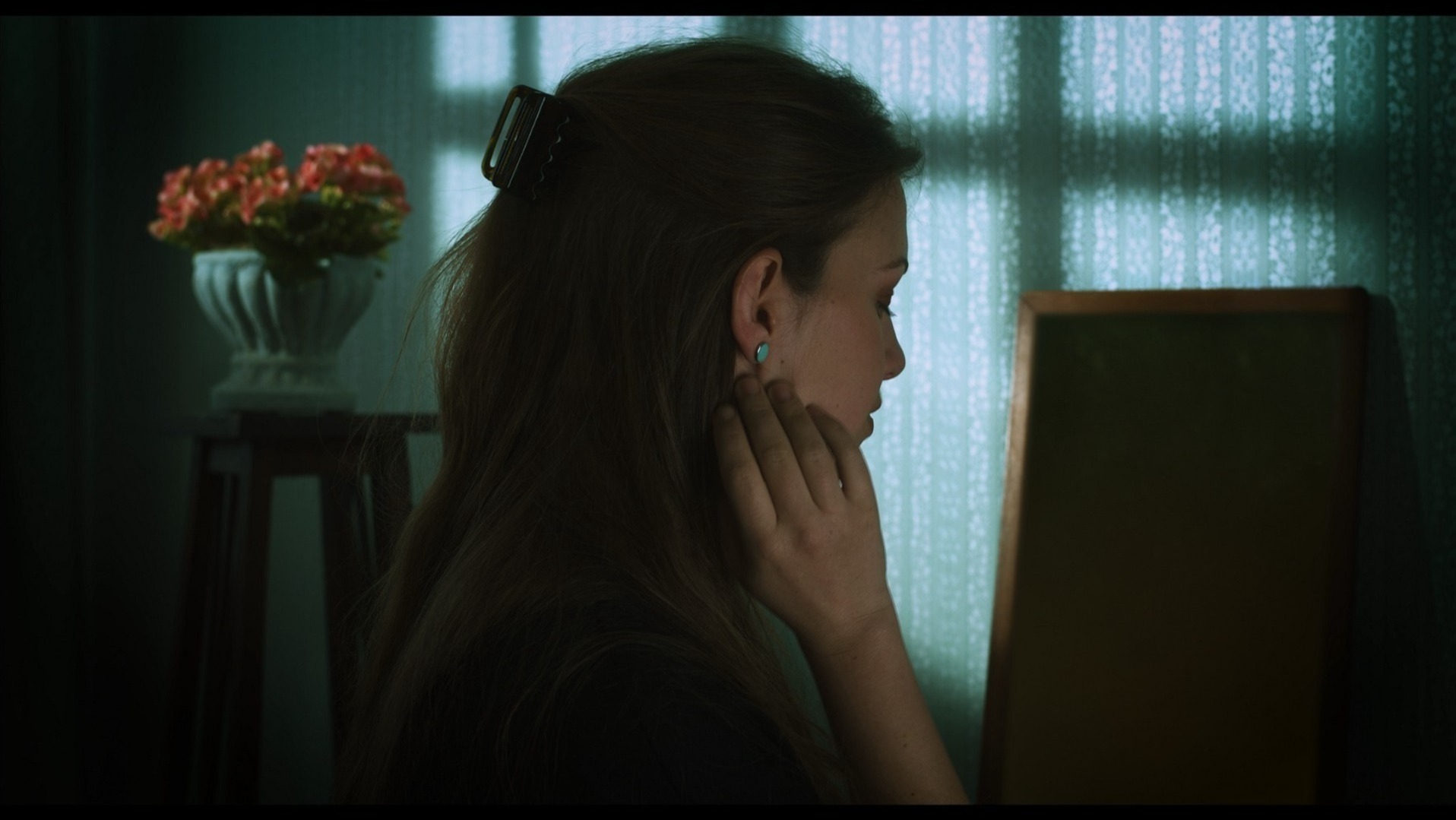 Profil kobiety z upiętymi włosami, dotykającej twarzy, w tle wazon z kwiatami i rama obrazu, oświetlona przez padające cienie na wzorzystej tapecie.