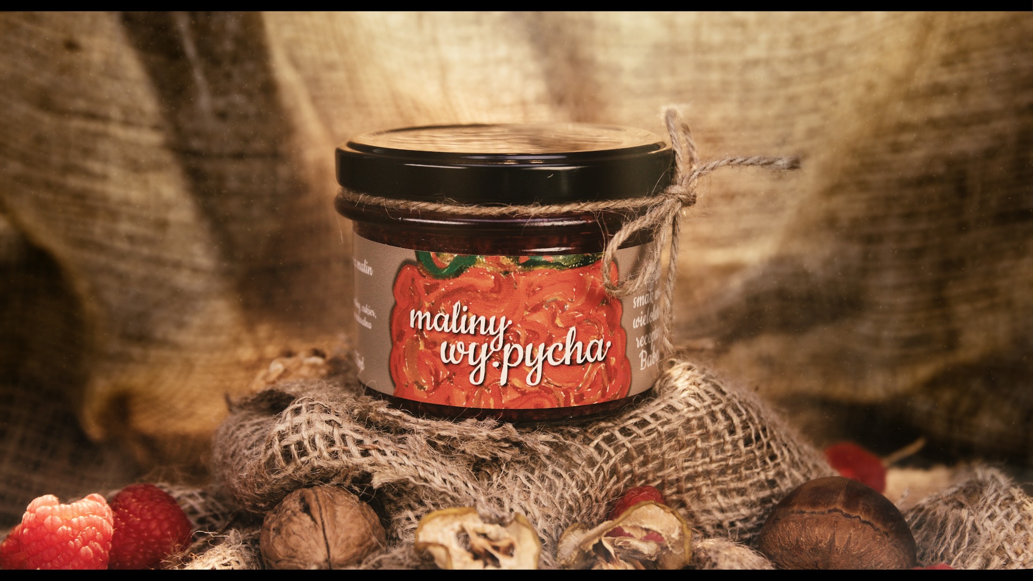 Słoik dżemu malinowego 'maliny wy-pycha' z etykietą ozdobioną rysunkiem malin, otoczony jutową tkaniną, orzechami włoskimi i świeżymi malinami, na tle drapowanej tkaniny.