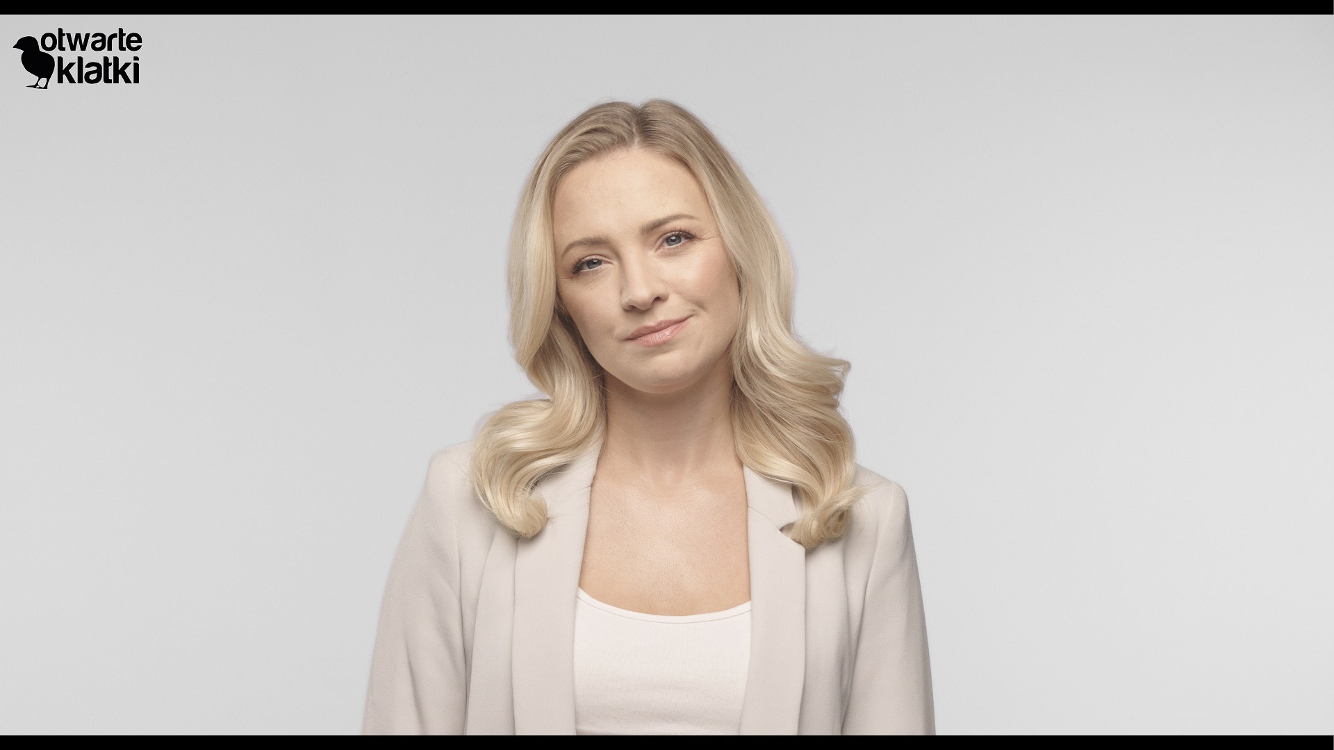 Portret kobiety z blond włosami w beżowej marynarce na jasnoszarym tle, z logo 'otwarte klatki' w lewym górnym rogu.