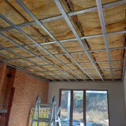 Montaż sufitu podwieszanego z wełną mineralną w pomieszczeniu z ceglaną ścianą i oknem, widoczne profile i materiały izolacyjne, drabina aluminiowa