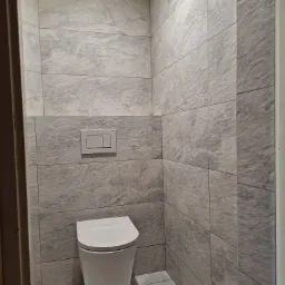 Nowoczesna toaleta podwieszana w łazience wykończonej szarymi płytkami imitującymi kamień, z widocznym przyciskiem spłukującym na ścianie i subtelnym oświetleniem.