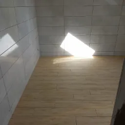 Świeżo wykafelkowane wnętrze z beżową podłogą imitującą drewno i szarymi, błyszczącymi płytkami na ścianach, oświetlone silnym, naturalnym światłem wpadającym przez okno.