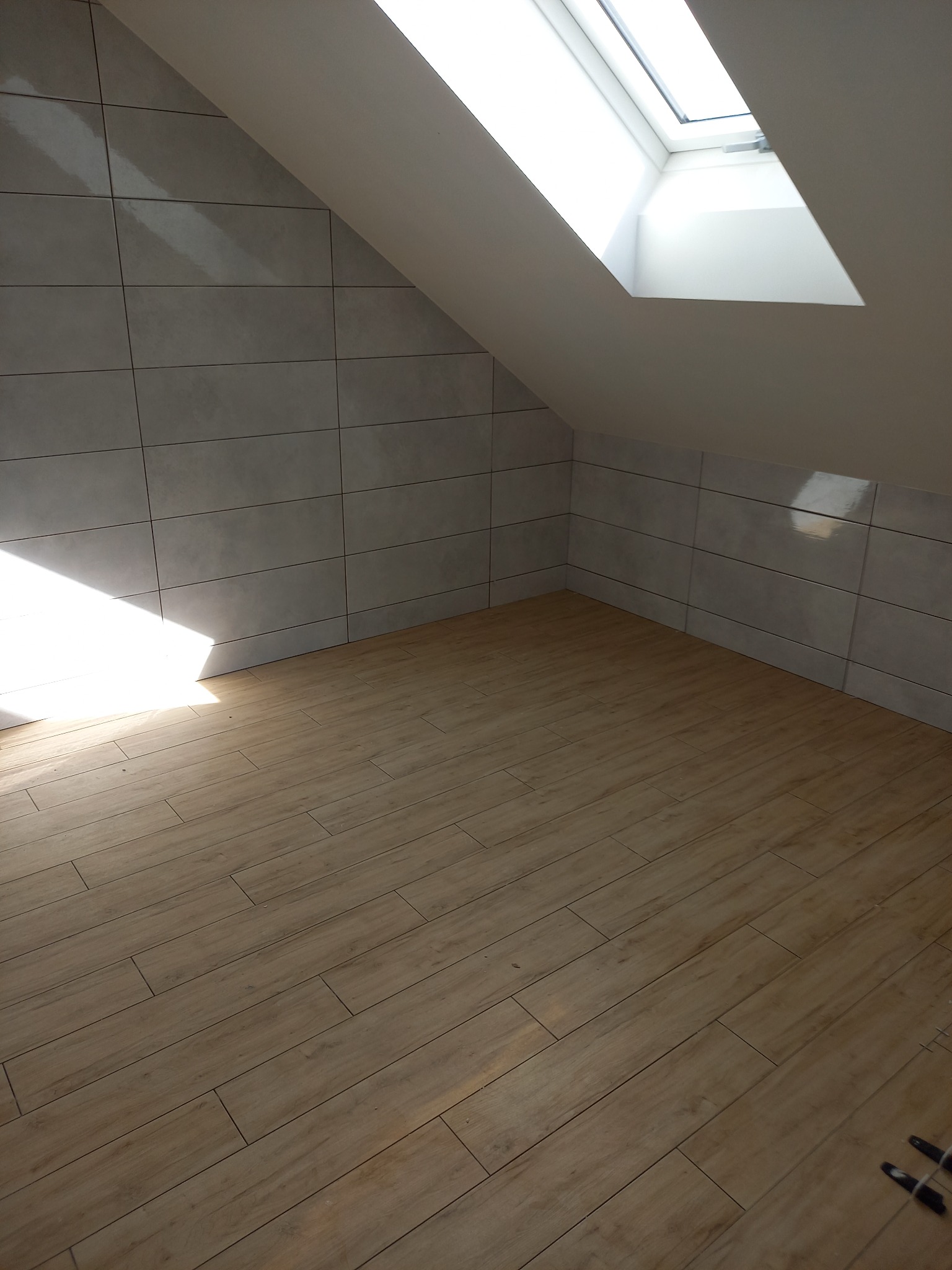 Poddasze po remoncie: szare płytki ceramiczne na ścianach i drewnopodobna podłoga z jasnym oknem dachowym.