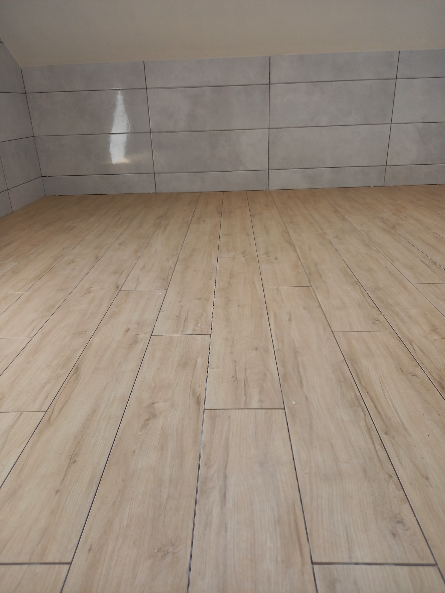 Ściana wyłożona szarymi, prostokątnymi płytkami ceramicznymi oraz podłoga wyłożona płytkami imitującymi drewno w jasnym odcieniu, ułożonymi wzdłużnie, w pomieszczeniu ze skosami.