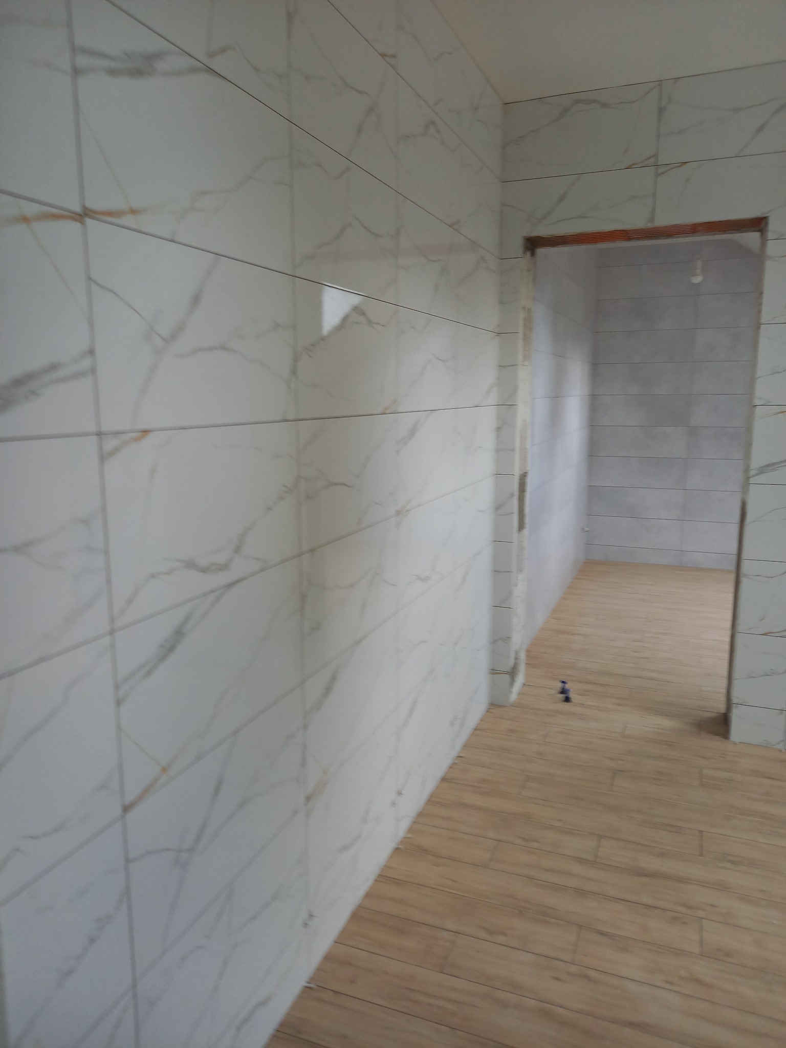 Ściana i podłoga wyłożone płytkami imitującymi marmur i drewno, widok na sąsiednie pomieszczenie przez surowe ościeżnice.