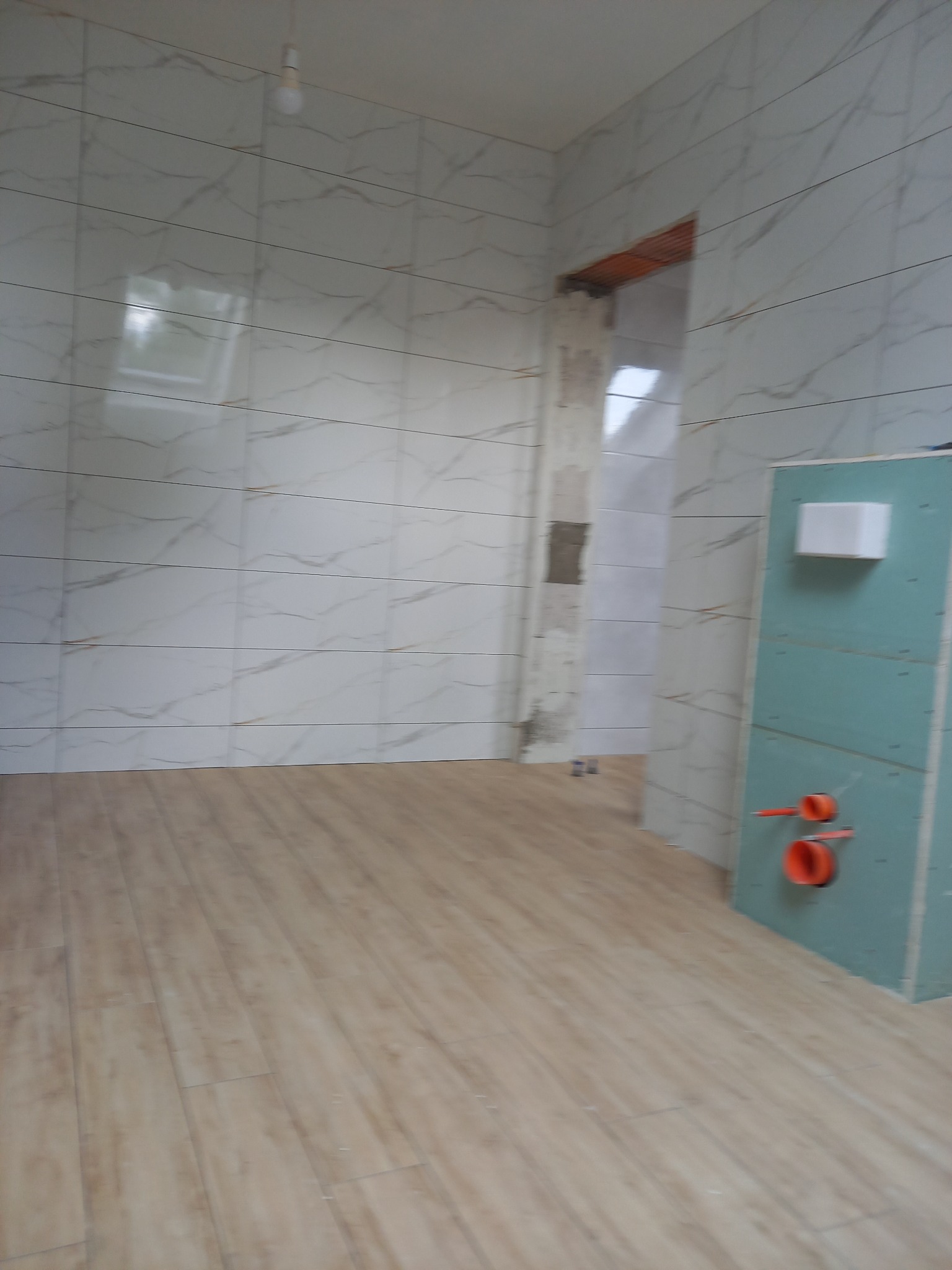 Wnętrze łazienki w trakcie prac wykończeniowych: marmurowe płytki ścienne, drewnopodobna podłoga, zabudowa z płyty karton-gips z widocznymi instalacjami hydraulicznymi, przygotowana do montażu...