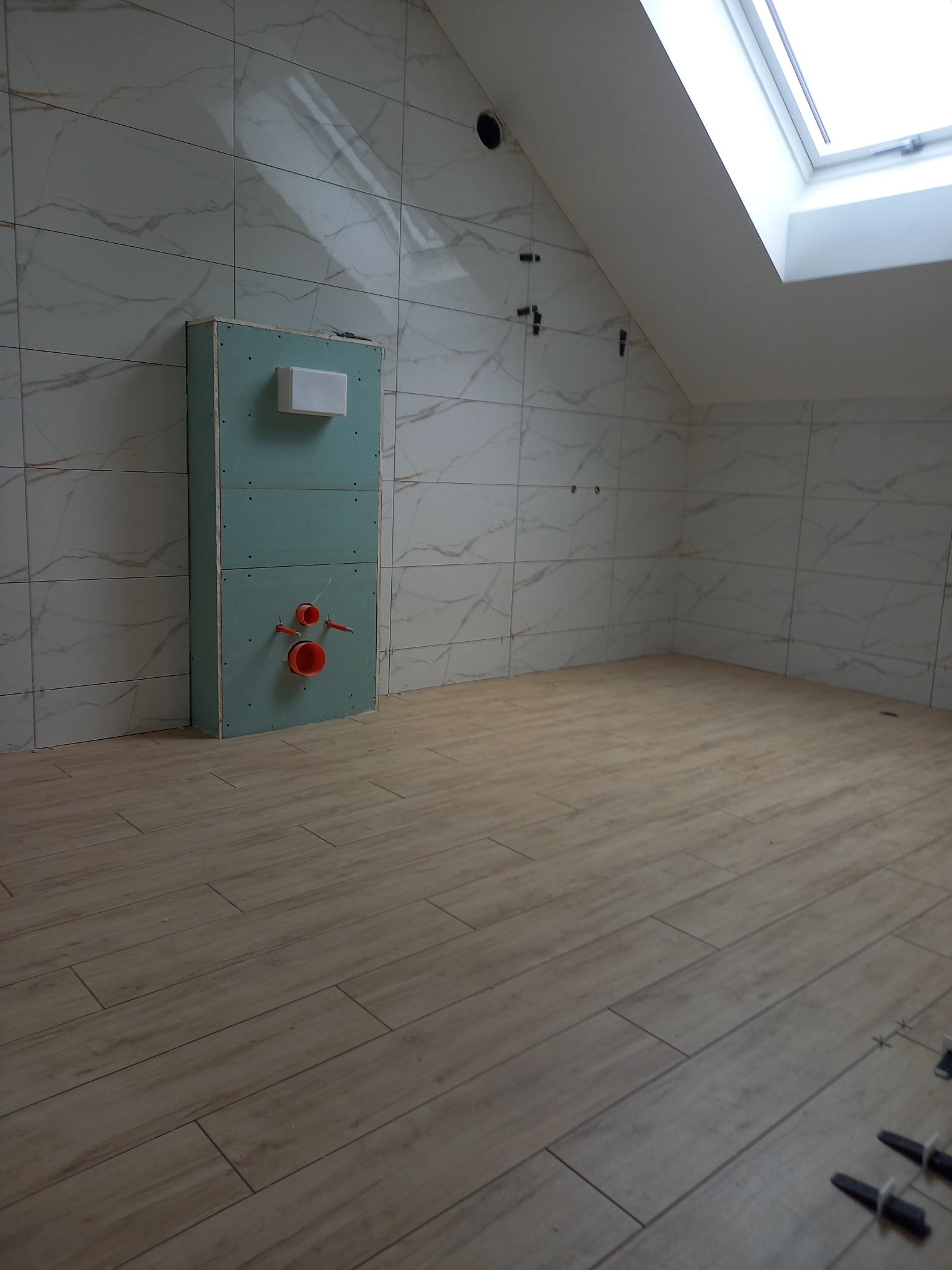 Łazienka na poddaszu w trakcie prac wykończeniowych: ułożone płytki imitujące drewno na podłodze, na ścianach białe płytki z marmurkowym wzorem, zabudowa stelaża WC z widocznymi przyłączami...