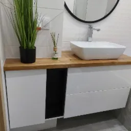 Biała szafka łazienkowa z drewnianym blatem, kwadratową umywalką i okrągłym lustrem na tle białych płytek, uzupełniona zieloną rośliną w doniczce i dyfuzorem zapachowym.