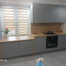 Nowoczesna kuchnia w odcieniach szarości z drewnianym blatem, zabudowa pod oknem z kwiatem i butelką, na blacie płyta indukcyjna i miska z owocami, na podłodze zabawka dziecięca.