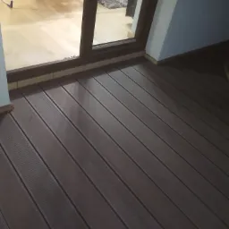 Ciemnobrązowe deski tarasowe kompozytowe ułożone na zewnątrz, widok z góry, z widocznym fragmentem wnętrza domu przez otwarte drzwi balkonowe.