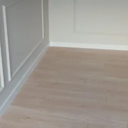 Jasne panele podłogowe ułożone w rogu pomieszczenia z dekoracyjnymi, białymi listwami przypodłogowymi i sztukaterią na ścianach.